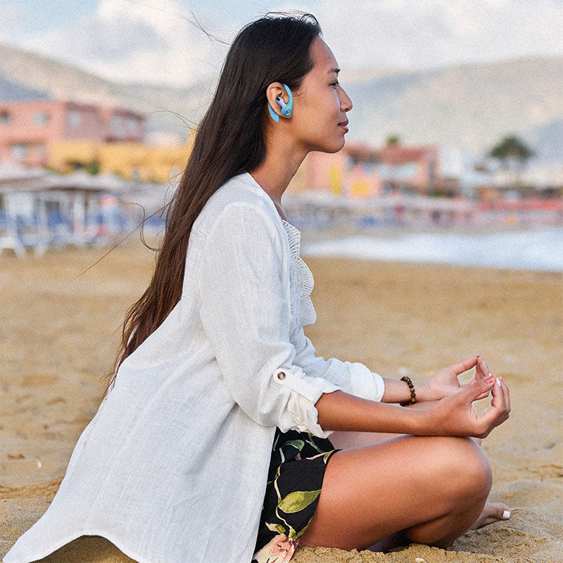 A woman peacefully meditating on the beach, during the day, while wearing baby blue AirPod pro ear hooks, an AirPod accessory.